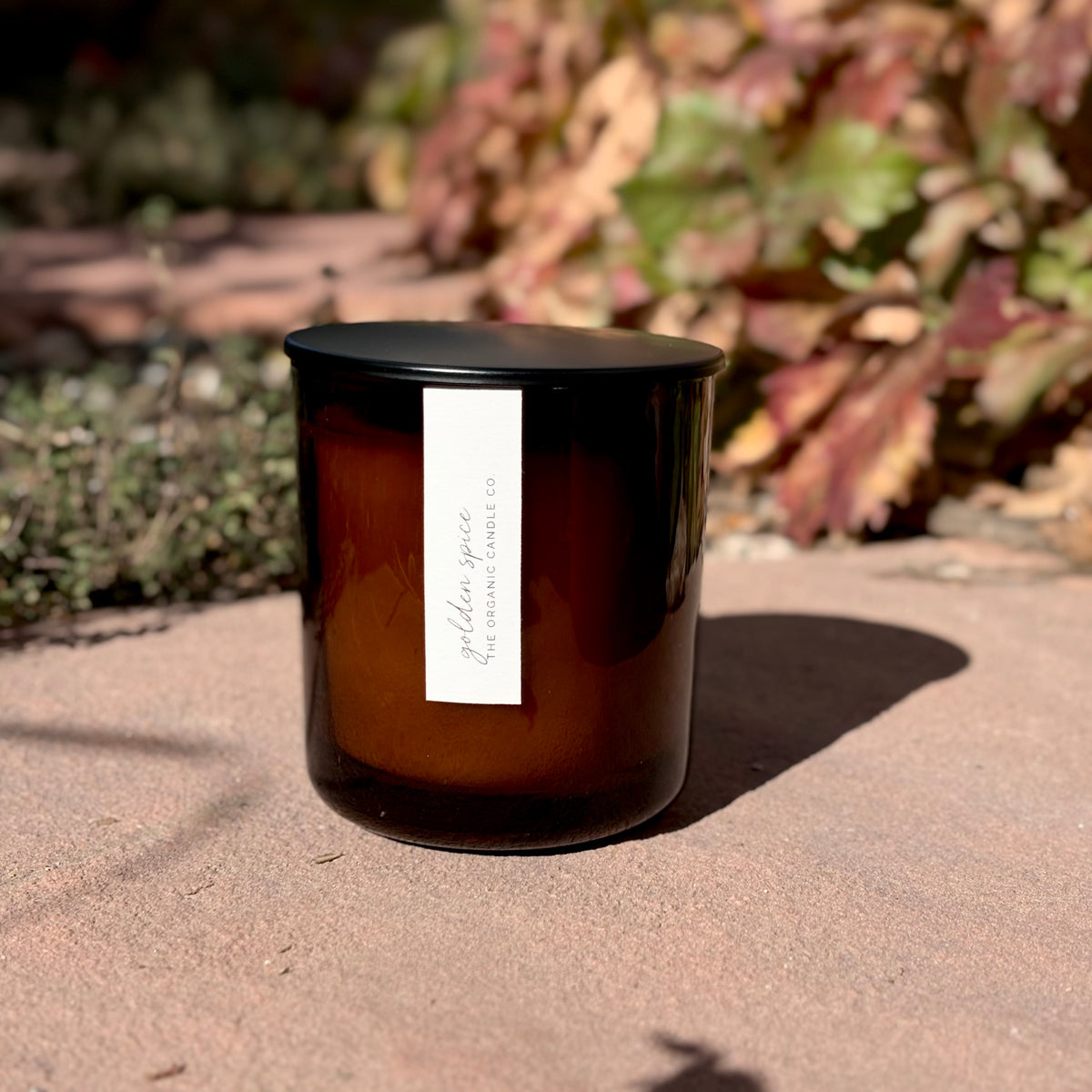 An organic candle in an amber glass jar candle with a black lid and a white vertical label reading 'THE ORGANIC CANDLE CO.', placed outdoors on a stone surface with blurred foliage in the background.