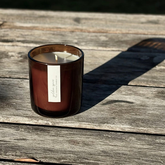 An organic candle in an amber glass jar candle with a white label sits on a weathered wooden surface in bright sunlight, casting a long shadow.