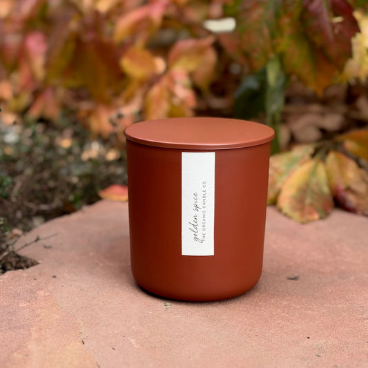 An organic candle in a matte terracotta-colored jar candle with a matching lid sits on a stone surface outdoors, with blurred autumn foliage in the background. A white vertical label on the front reads 'golden spice' and 'The Organic Candle Co.