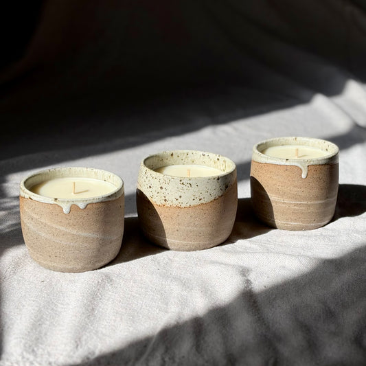 Three organic candles in handmade ceramic cups with a speckled beige and brown glaze and a contrasting cream-colored dripping rim, set on a textured light gray surface under dramatic natural sunlight and shadows.