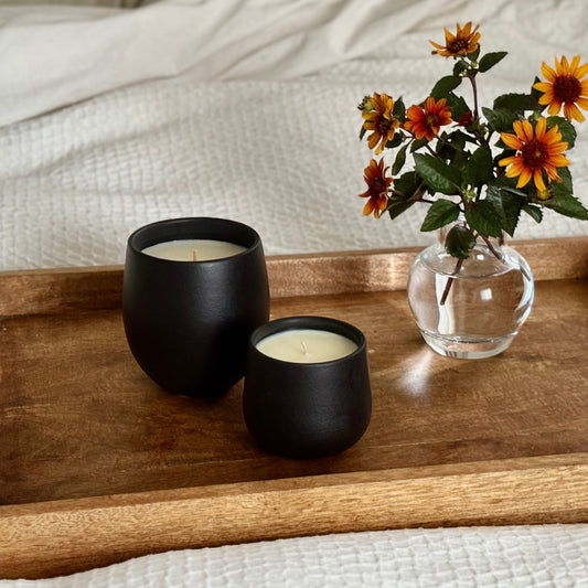 Two black ceramic organic candles, one large and one small, on a wooden tray that is resting on a white comforter. Flowers are off to the side.