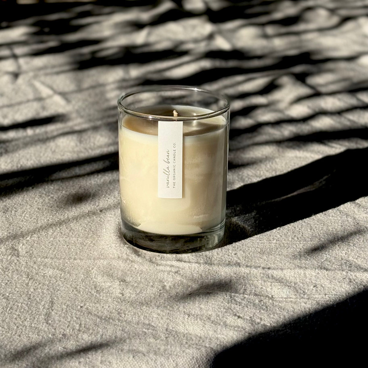 An organic candle in a clear glass jar sits on a textured beige fabric surface, lit by natural sunlight with dappled shadows cast across the scene. A small white label on the front reads 'vanilla bean' and 'THE ORGANIC CANDLE CO.