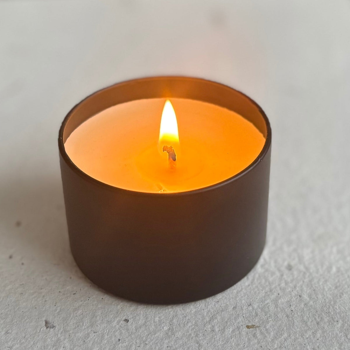 Organic candle in a black cylindrical container with a lit flame on a neutral background