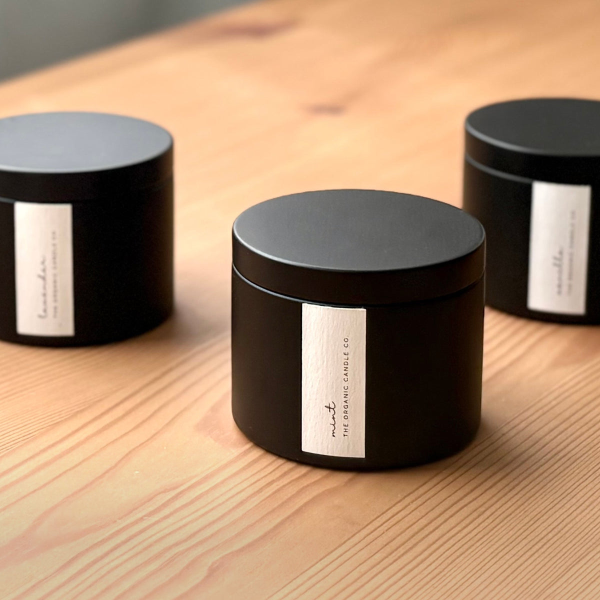 Three black candles with labels on a wooden surface, featuring 'The Organic Candle Co'.