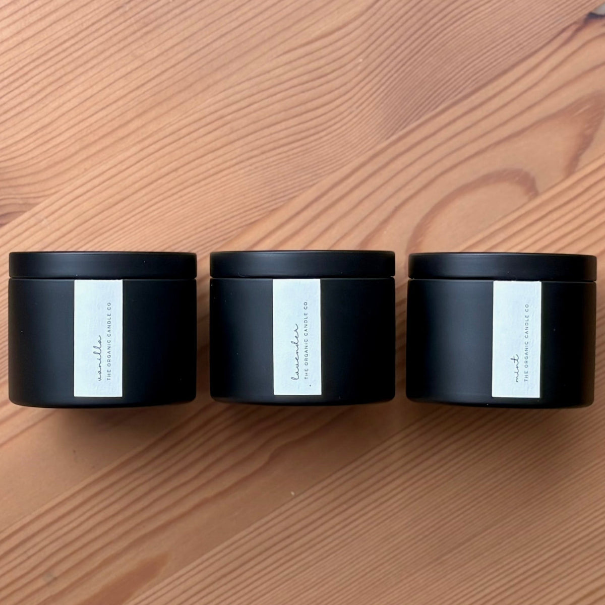 Three organic candles in black cylindrical containers white labels on a wooden surface, branded 'The Organic Candle Co'.
