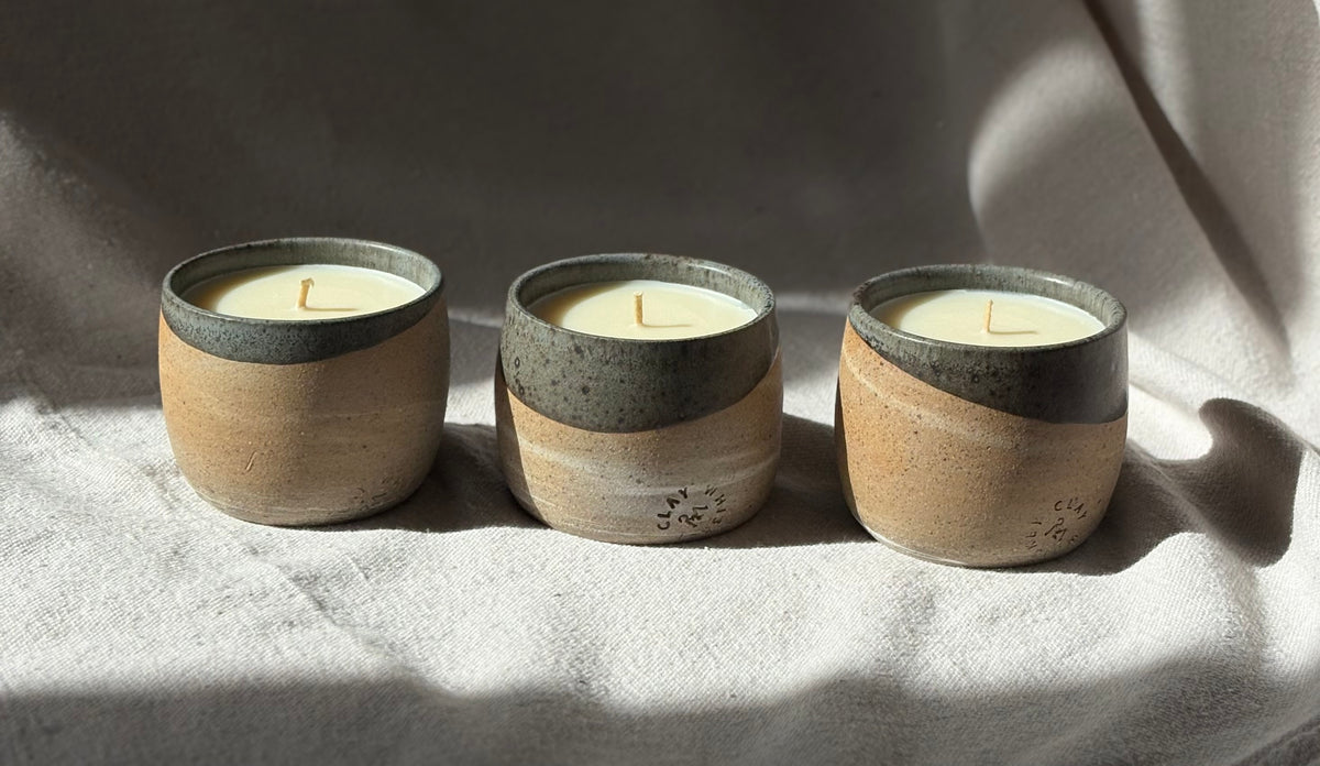 Three organic candles in ceramics with a speckled dark gray top and a warm beige lower body, arranged side by side on a draped light fabric under direct sunlight.