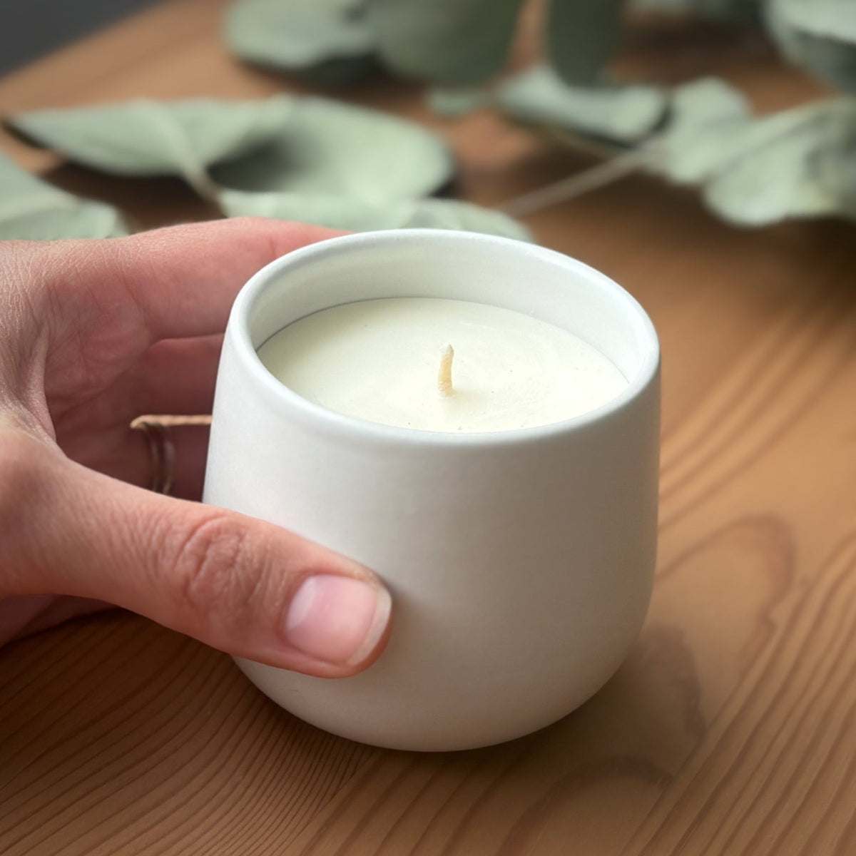 Hand holding a white organic candle on a wooden surface.