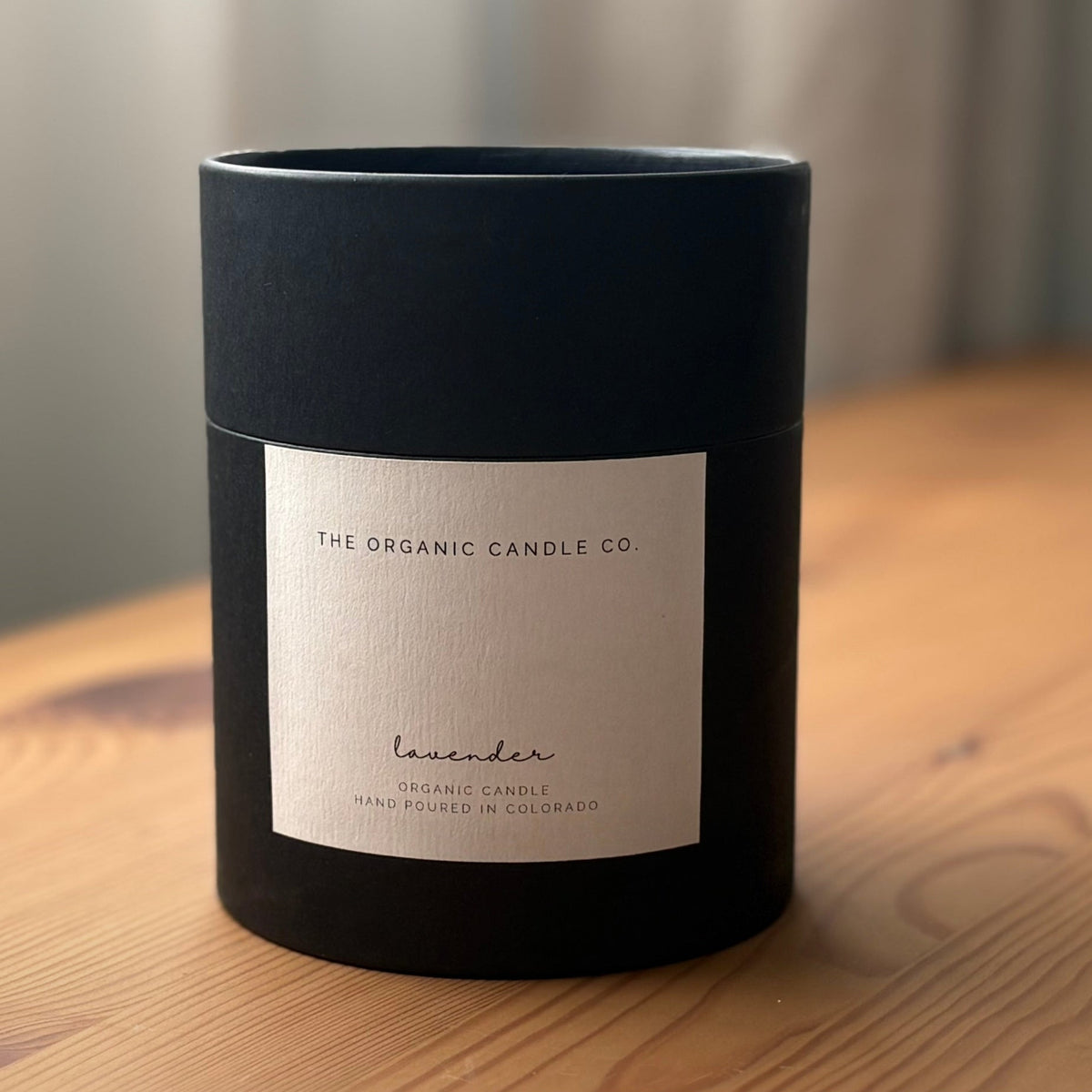 Black cylindrical container with a label on a wooden surface, featuring 'The Organic Candle Co.'