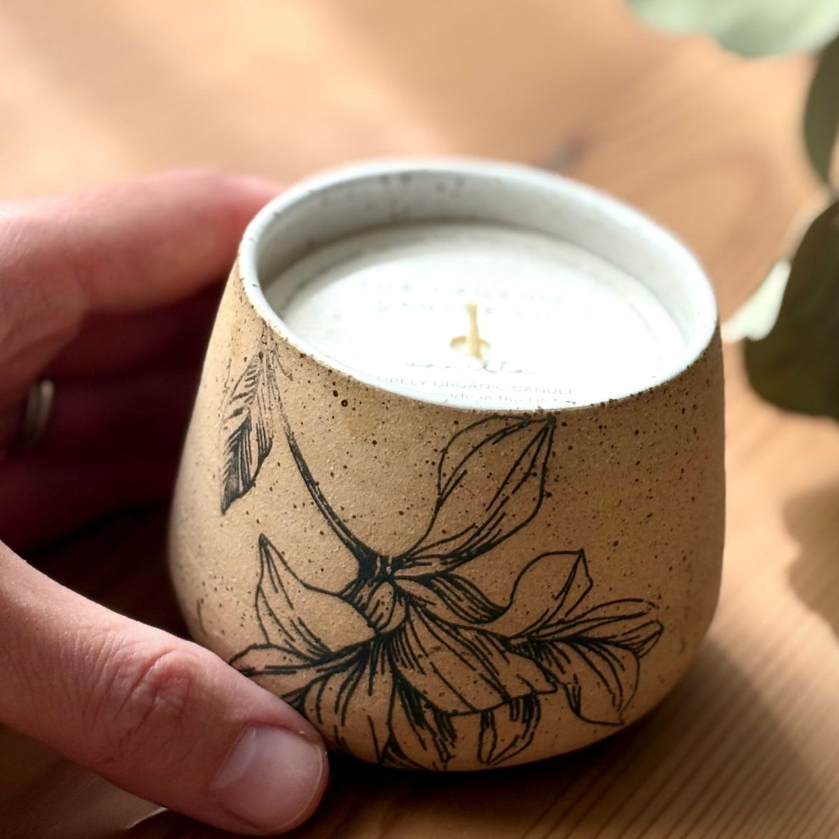Organic candle in ceramic vessel with botanical design, held by a hand on a wooden surface.