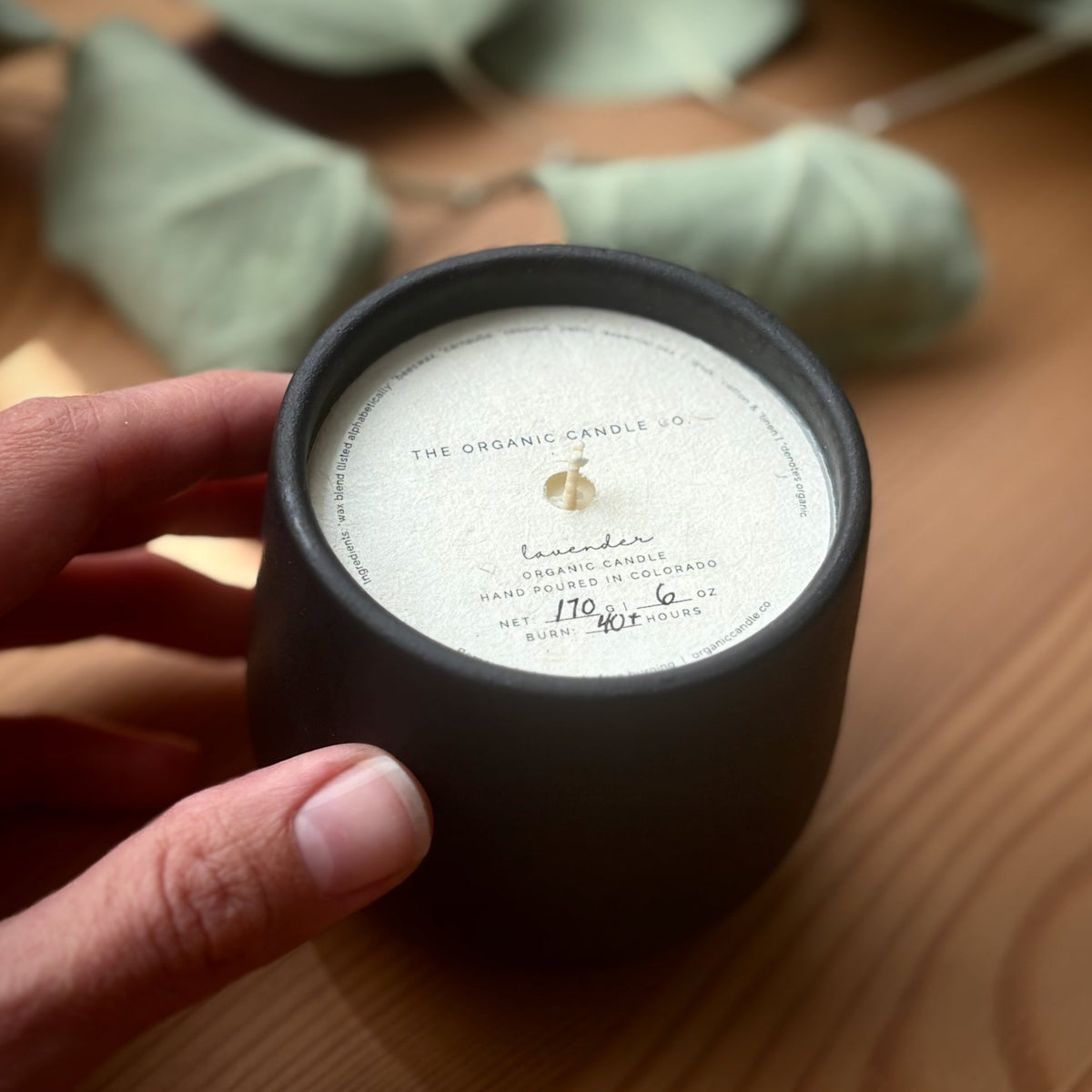Organic candle in black ceramic vessel held by a hand on a wooden surface, featuring 'The Organic Candle Co.'