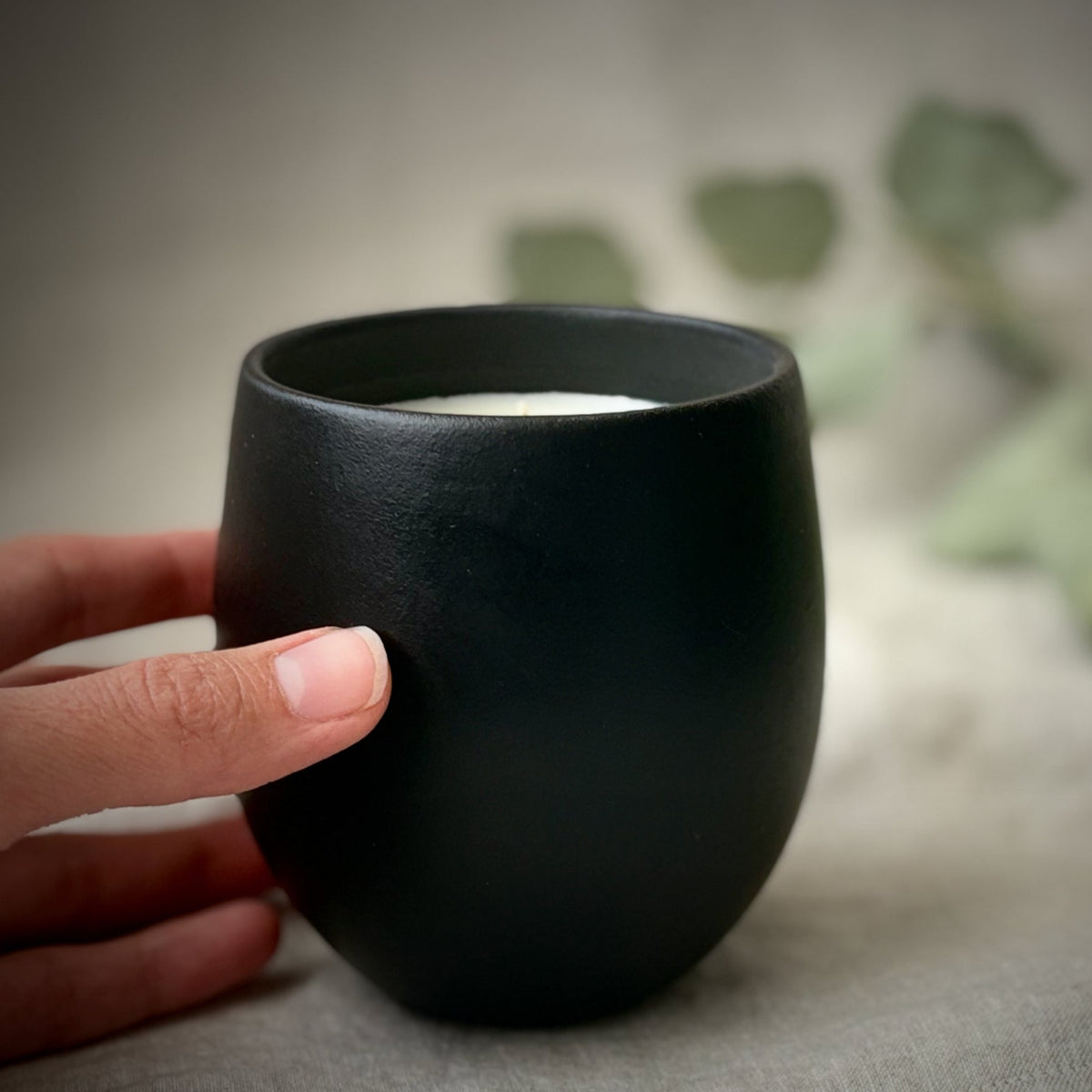 Black organic candle with a hand holding it, blurred greenery in the background, 'The Organic Candle Co.' branding.