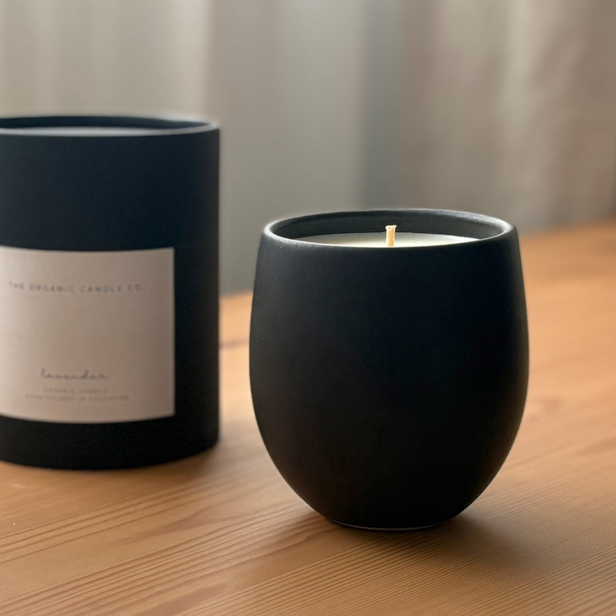 Organic candle in black ceramic vessel and black cylindrical packaging on a wooden surface, branded 'The Organic Candle Co'.