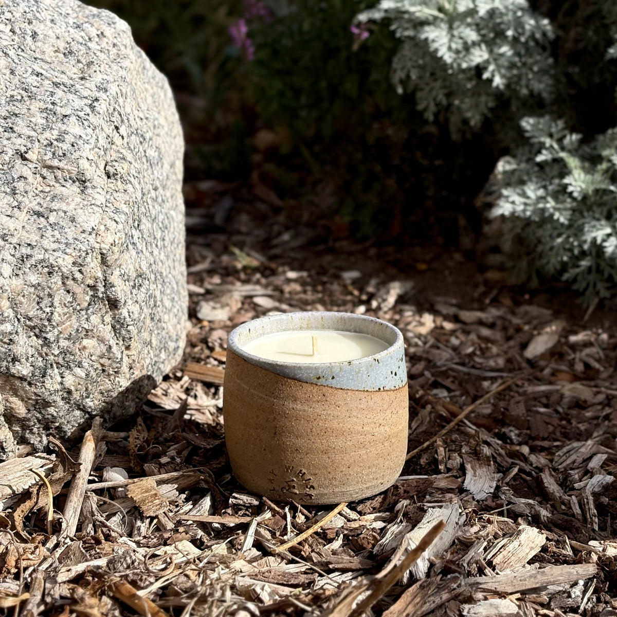 An organic candle in a ceramic cup with a speckled light blue rim and a warm tan body sits on a bed of wood chips outdoors next to a large rock and greenery.