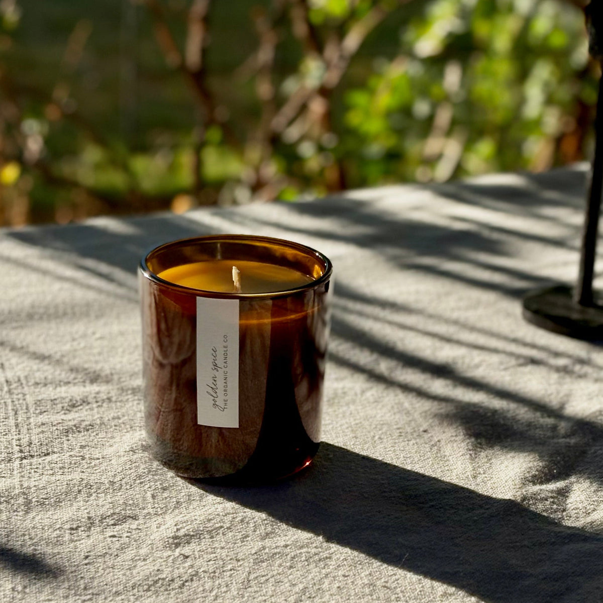 An organic candle in an amber glass jar with a white label sits on a textured gray linen cloth outdoors, with dappled sunlight and a blurred green garden background.