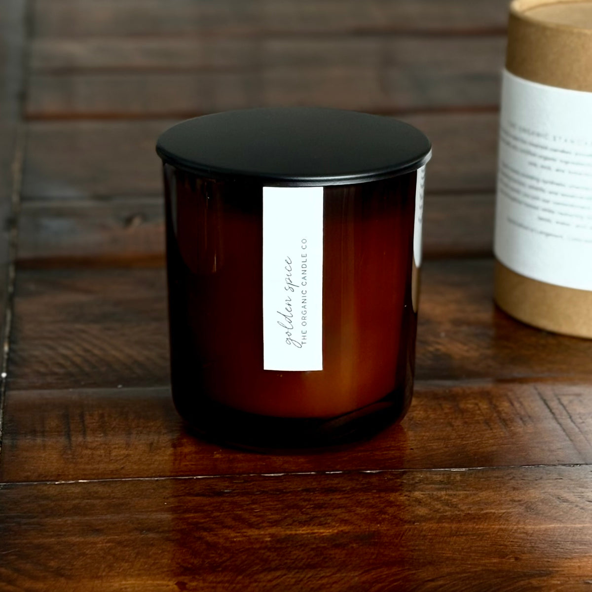 An organic candle in an amber glass jar with a black lid and a white vertical label reading 'Golden Spice' and 'The Organic Candle Co.' on a dark wooden surface, with a kraft cylinder box partially visible in the background.