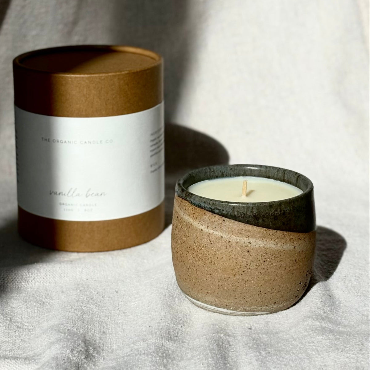 An organic candle in a speckled beige ceramic with a dark glazed rim sits next to its brown kraft paper packaging on a draped neutral fabric background.
