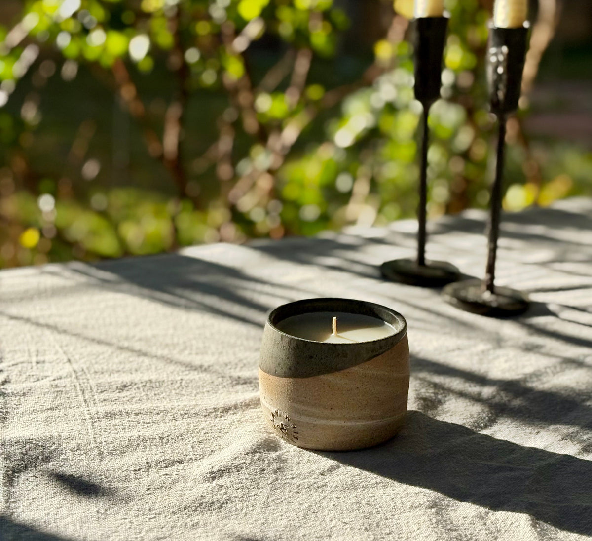 An organic candle in a handmade ceramic with a two-tone beige and dark gray glaze sits on a textured light gray cloth outdoors, with blurred greenery in the background and candlesticks behind it.