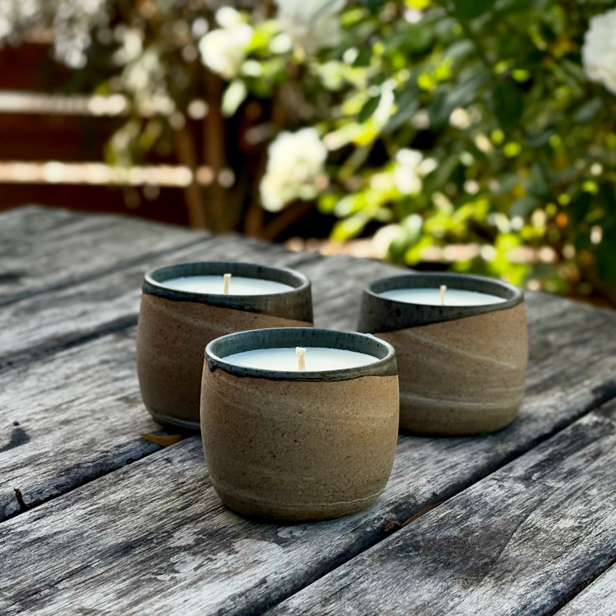 Three small organic candles in textured earthy ceramic cups are arranged on a weathered wooden table outdoors, with blurred greenery and white flowers in the background.