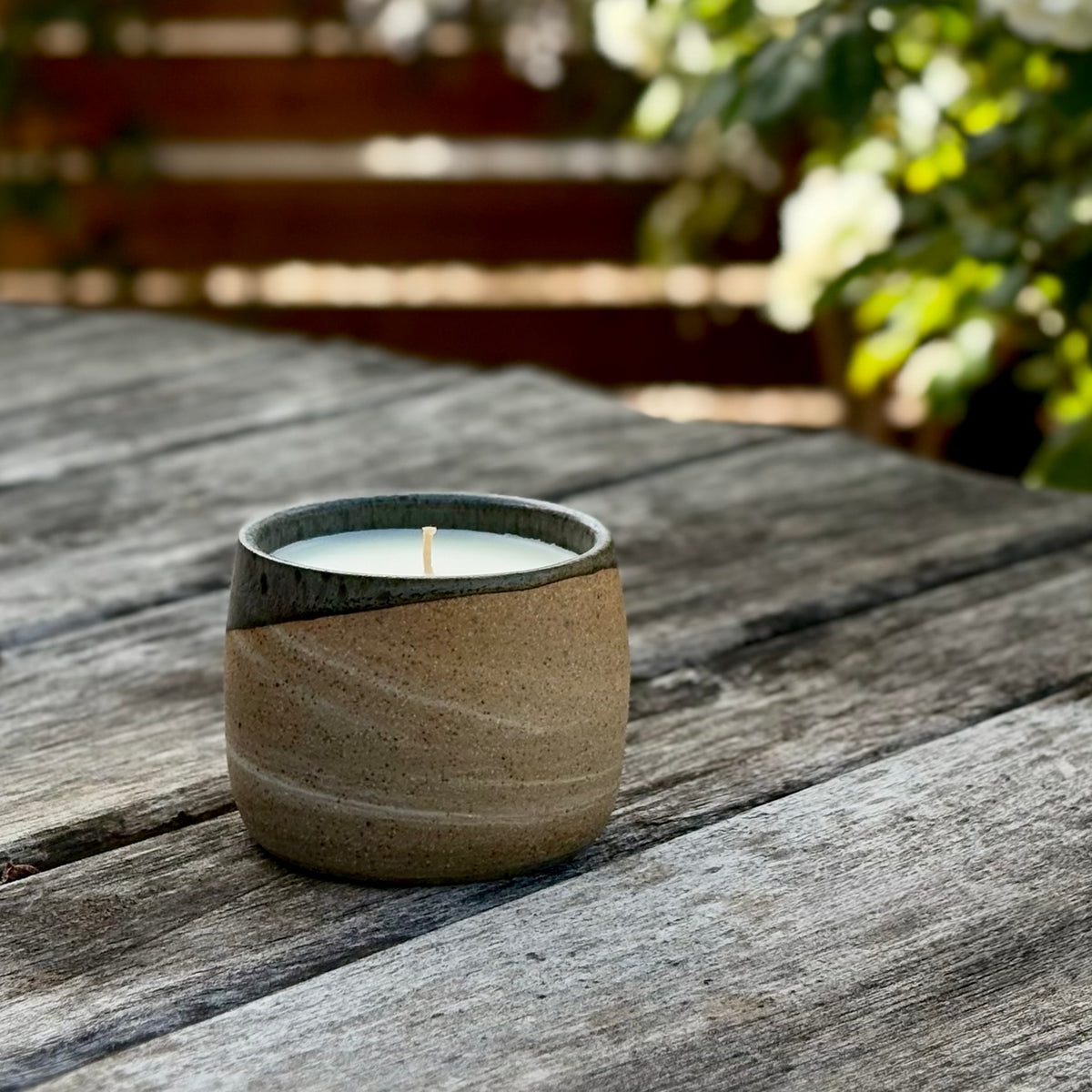 A small organic candle in a speckled ceramic beige cup with a dark gray rim sits on a weathered wooden table outdoors, with blurred green foliage in the background.