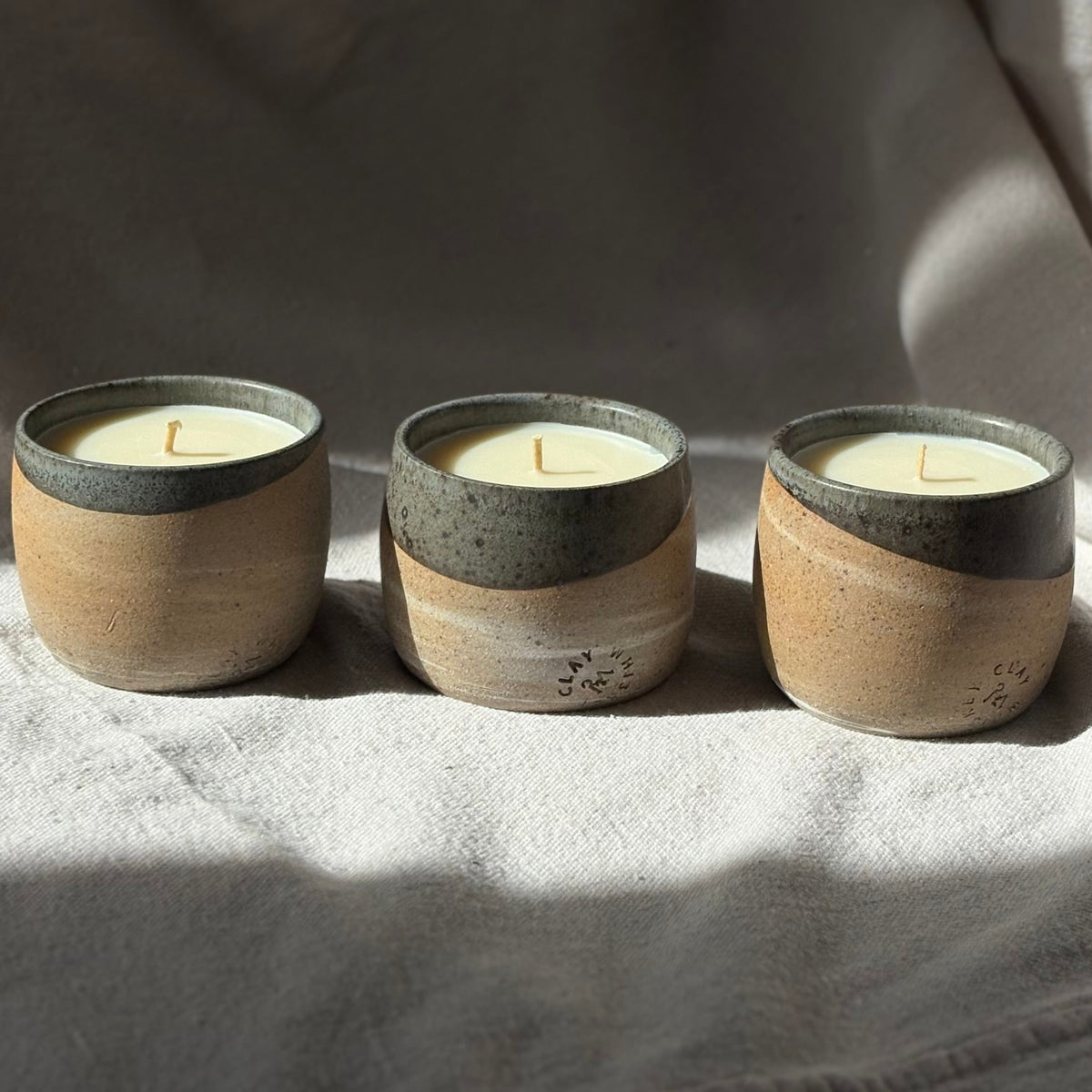 Three small ceramic organic candles in handmade speckled stoneware cups with a two-tone design, sitting on a textured light gray fabric under natural sunlight.