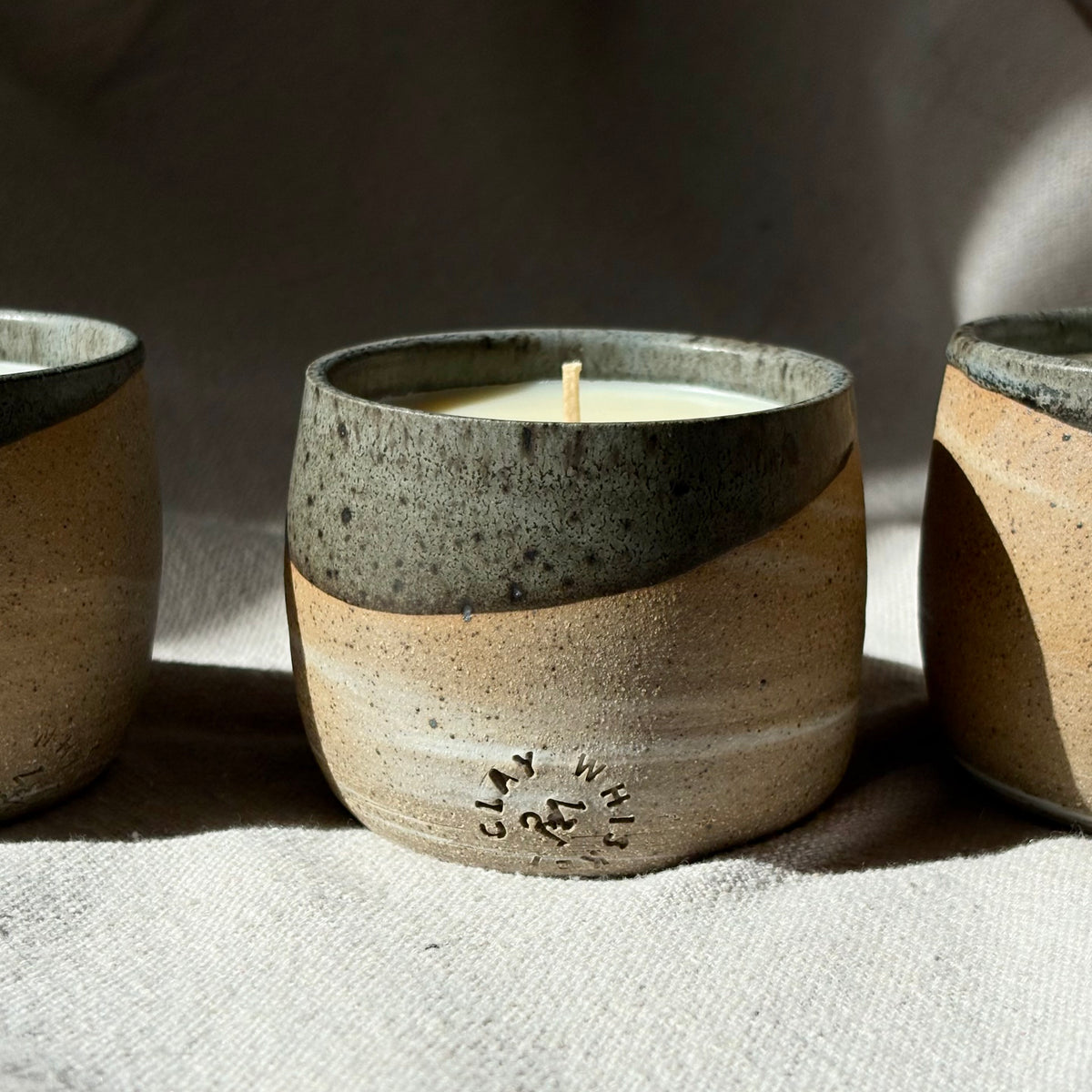 An organic candle in a handmade ceramic with a two-tone design, featuring a speckled dark gray top half and a sandy beige textured bottom half. The candle is styled on a neutral fabric surface with soft, natural shadows.