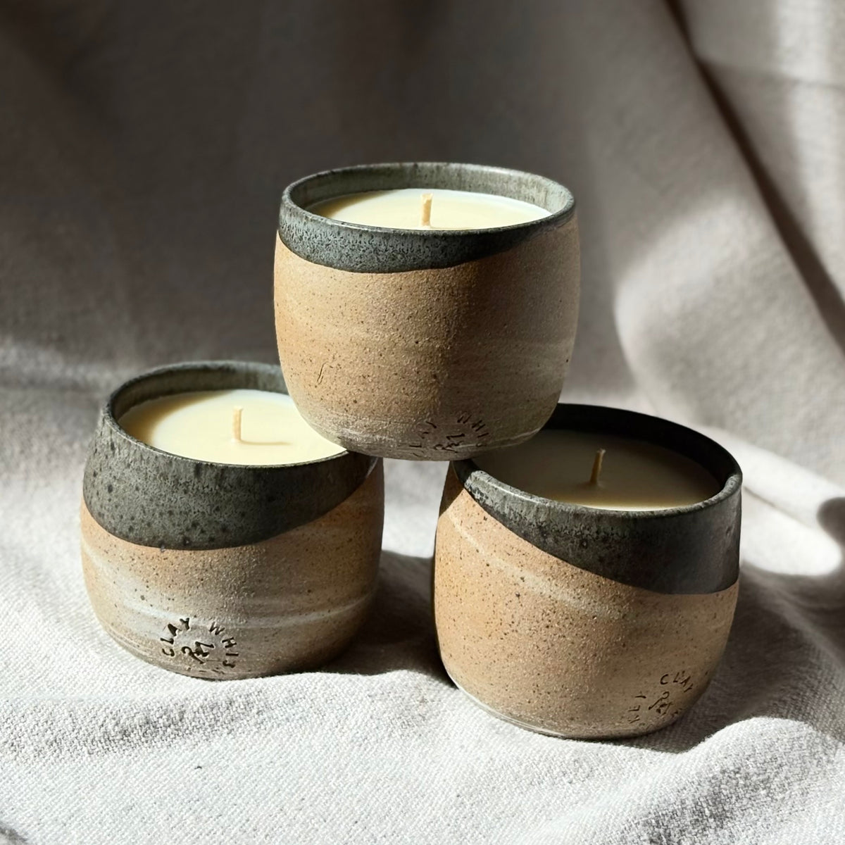 Three small organic candles in ceramic cups with a speckled beige body and a dark charcoal-gray rim, arranged in a pyramid on draped light beige fabric.