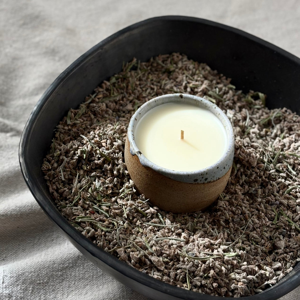 A small organic candle in a ceramic cup sits in the center of a larger black bowl filled with dried lavender buds, on a neutral linen fabric background.