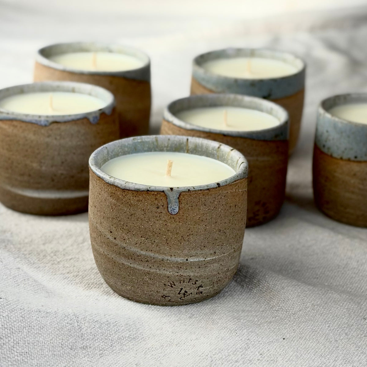 Six organic candles in handmade speckled brown and purple ceramic cups, arranged on a textured beige linen fabric background.