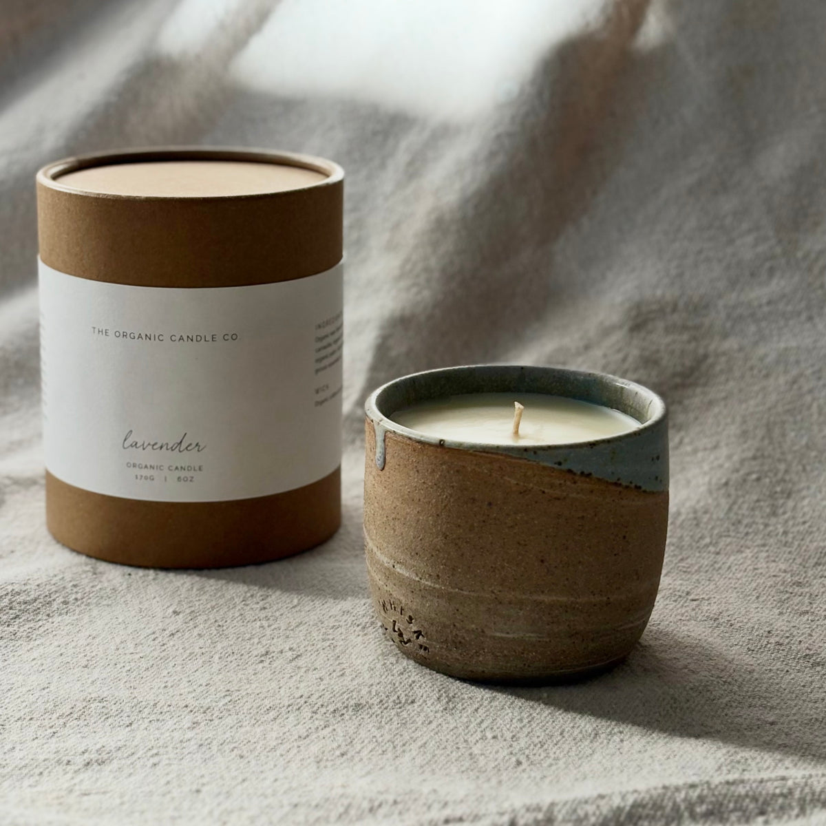 An organic candle in a ceramic cup and its kraft paper packaging box on a textured neutral fabric background. The candle has a rustic brown speckled ceramic body with a light blue glazed rim.
