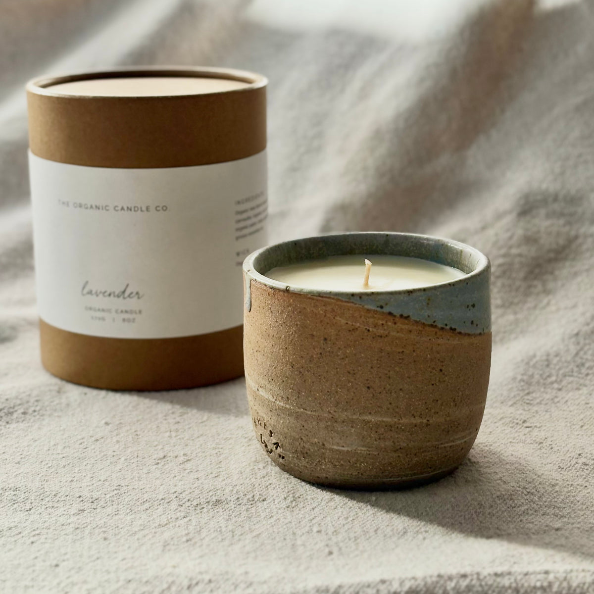 An organic candle in a ceramic cup and its kraft paper packaging box on a textured neutral fabric background. The candle has a rustic brown speckled ceramic body with a light blue glazed rim.