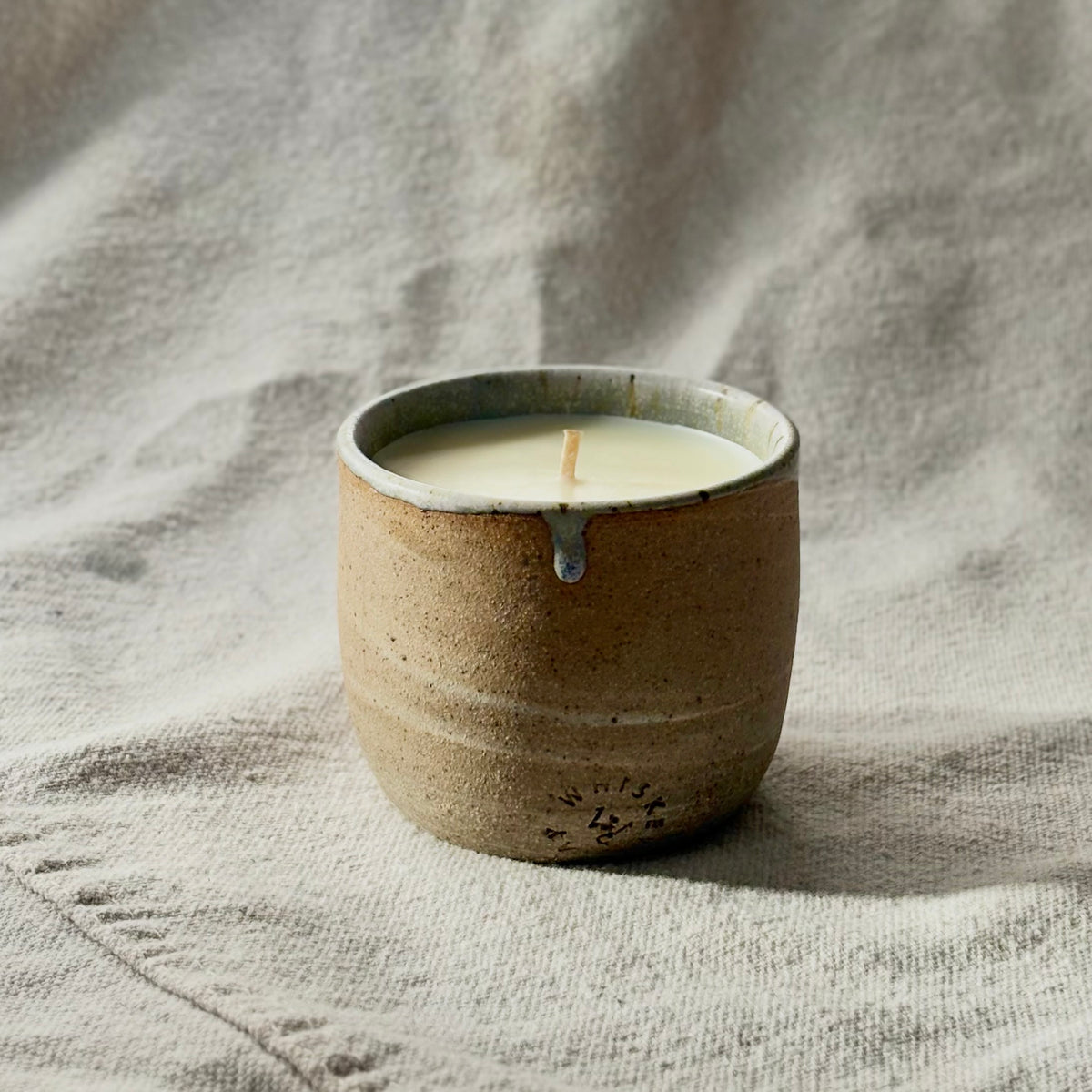 An organic candle in a ceramic cup on a textured neutral fabric background. The candle has a rustic brown speckled ceramic body with a light blue glazed rim.