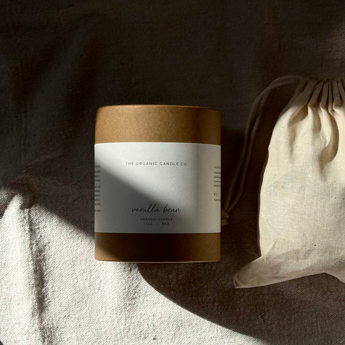 A kraft paper candle jar labeled 'THE ORGANIC CANDLE CO vanilla bean' sits on textured fabric next to a small off-white drawstring pouch in soft, directional sunlight.