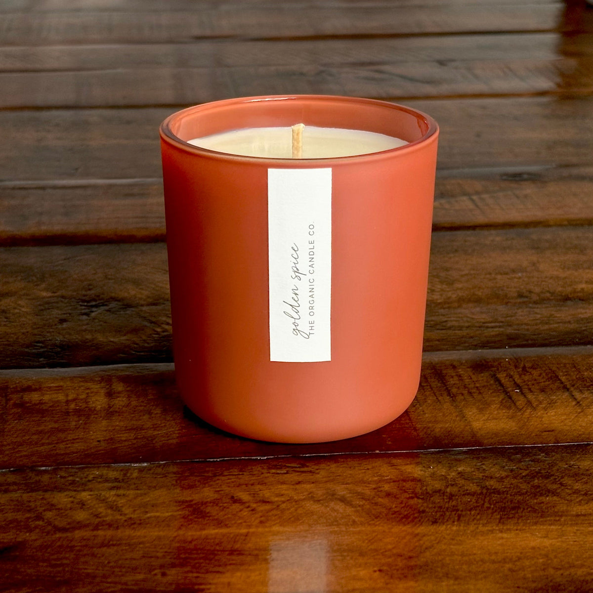 An organic candle in a matte terracotta-colored jar candle sits on a dark wooden table. A white vertical label on the front reads 'golden spice' and 'GOLDEN SPICE ORGANIC CANDLE CO.
