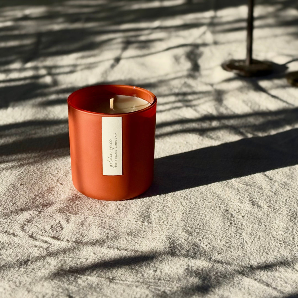 An organic candle in a matte orange-red candle glass jar sits on textured fabric outdoors, with dappled sunlight and shadows cast across the scene. A small white label is attached to the front with 'golden spice' and 'THE ORGANIC CANDLE CO.'