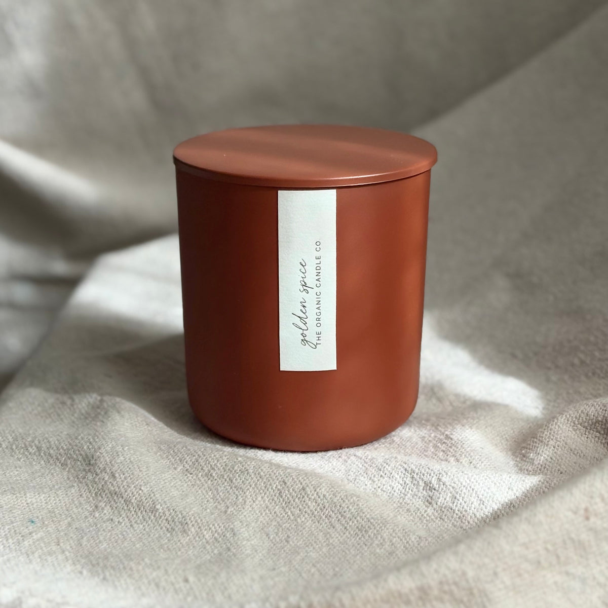 An organic candle in a matte terracotta-colored candle jar with a matching lid and a vertical white label reading 'golden spice' and 'The Organic Candle Co.', set on a textured beige fabric background.