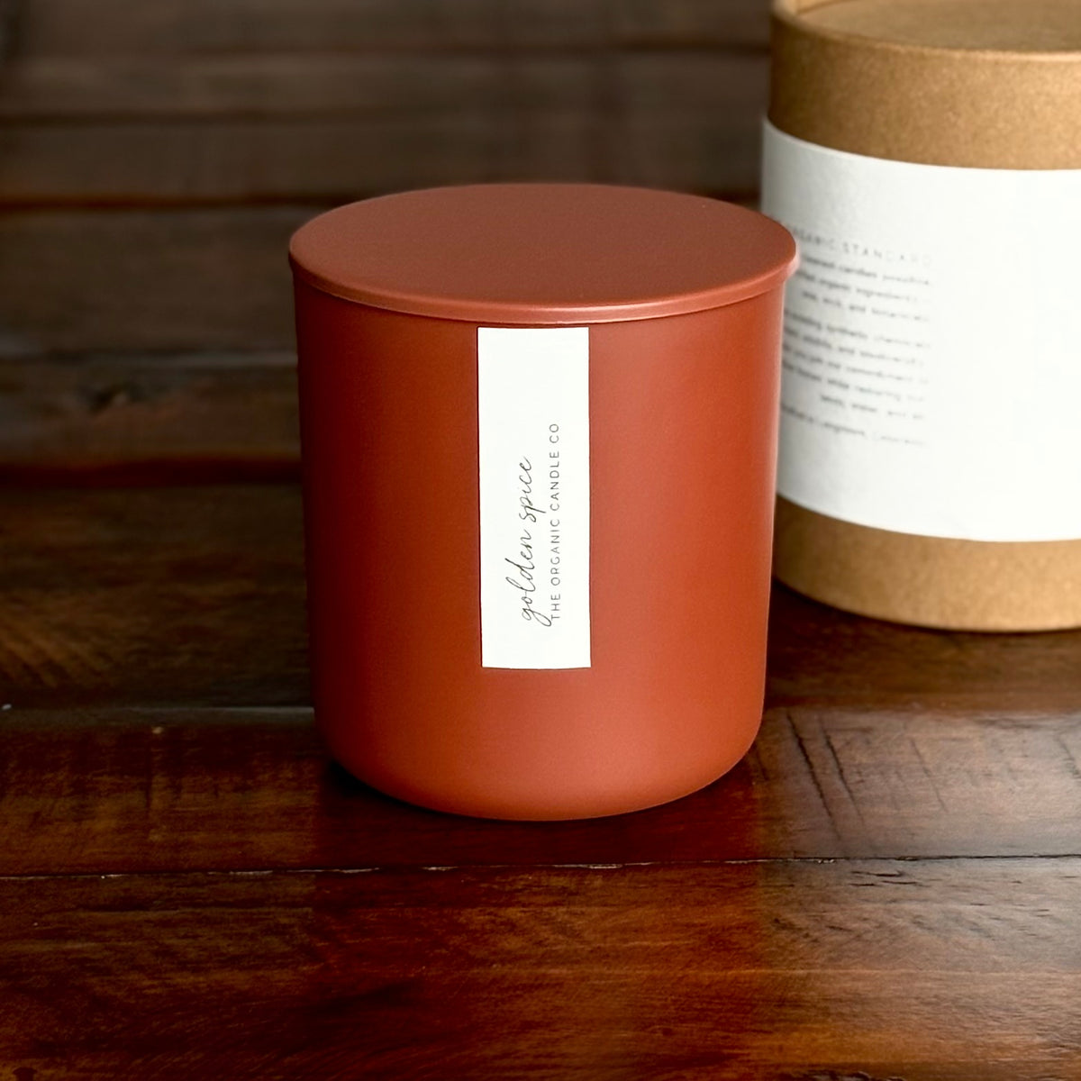 An organic candle in a matte terracotta-colored jar candle with a matching lid sits on a dark wooden table. A white vertical label on the front reads 'golden spice' and 'GOLDEN SPICE ORGANIC CANDLE CO.