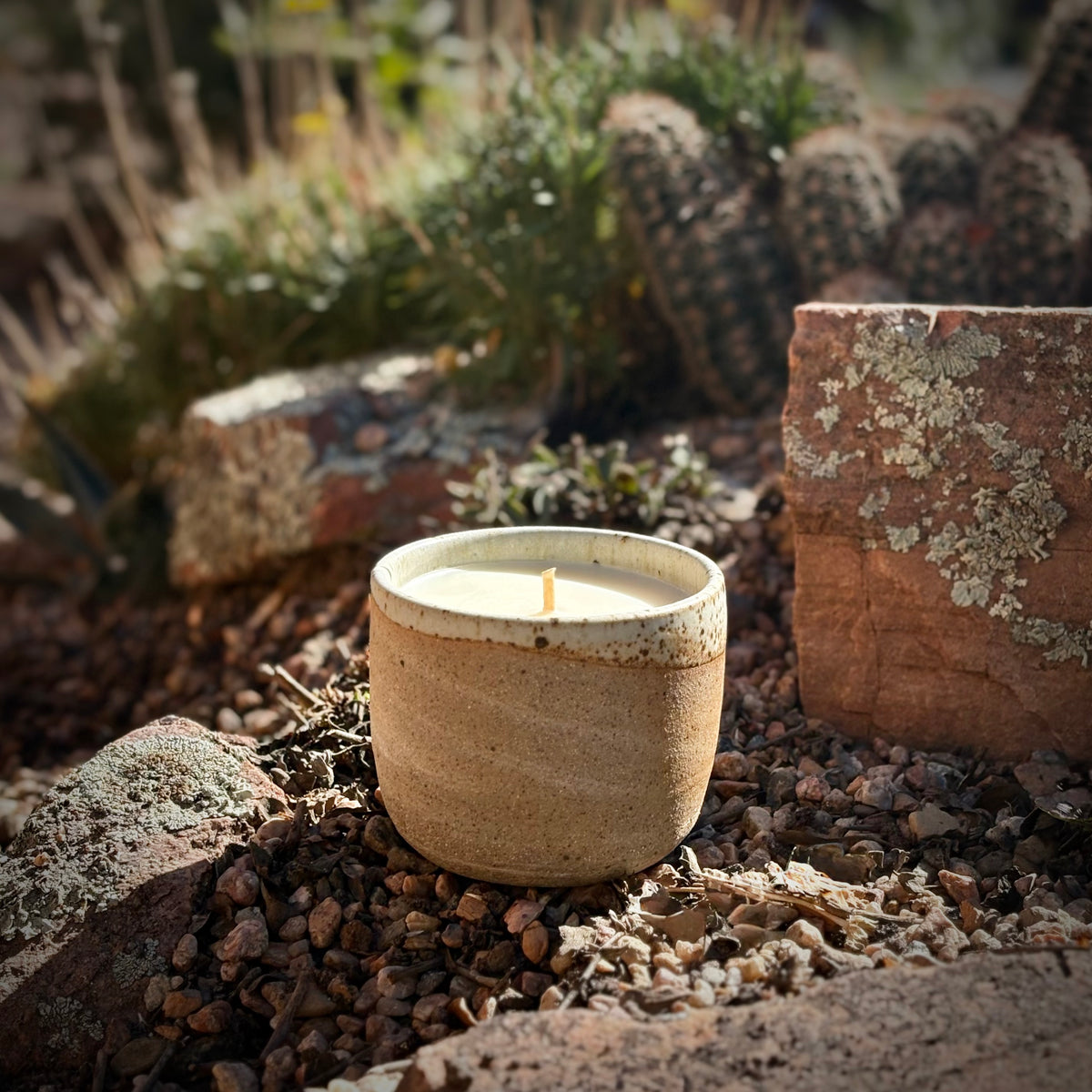 A small unlit organic candle in a handmade, speckled beige ceramic pot sits outdoors on a bed of small brown pebbles, surrounded by rocks, dried leaves, and blurred cacti in the background.