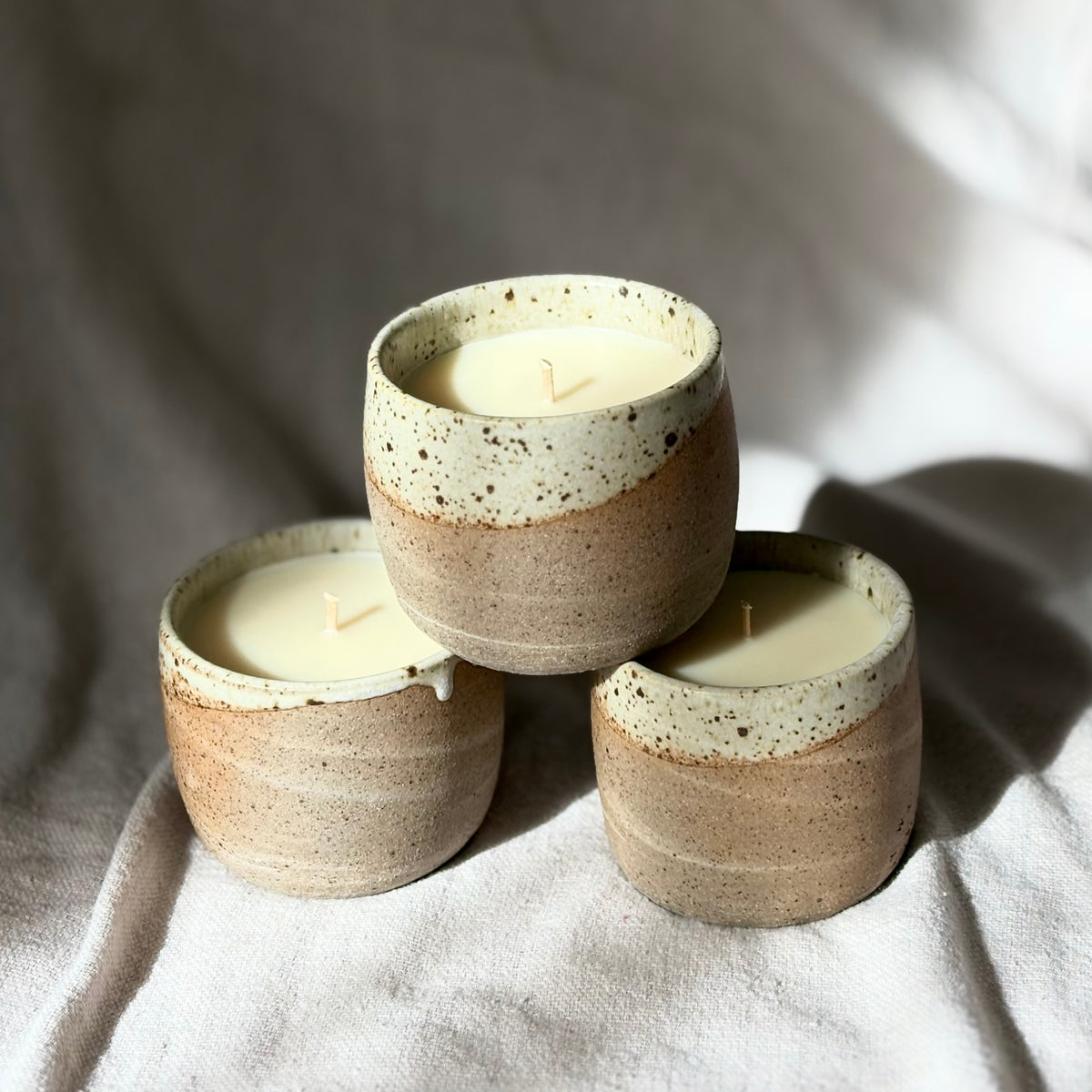 Three organic candles in handmade ceramic cups with a speckled beige and brown glaze and a contrasting cream-colored dripping rim, set on a textured light gray surface under dramatic natural sunlight and shadows.