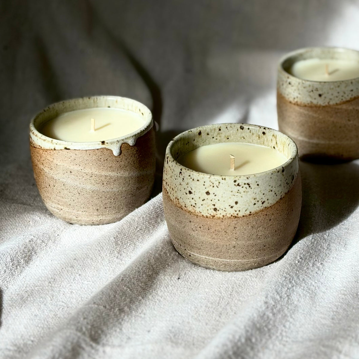 Three organic candles in handmade ceramic cups with a speckled beige and brown glaze and a contrasting cream-colored dripping rim, set on a textured light gray surface under dramatic natural sunlight and shadows.