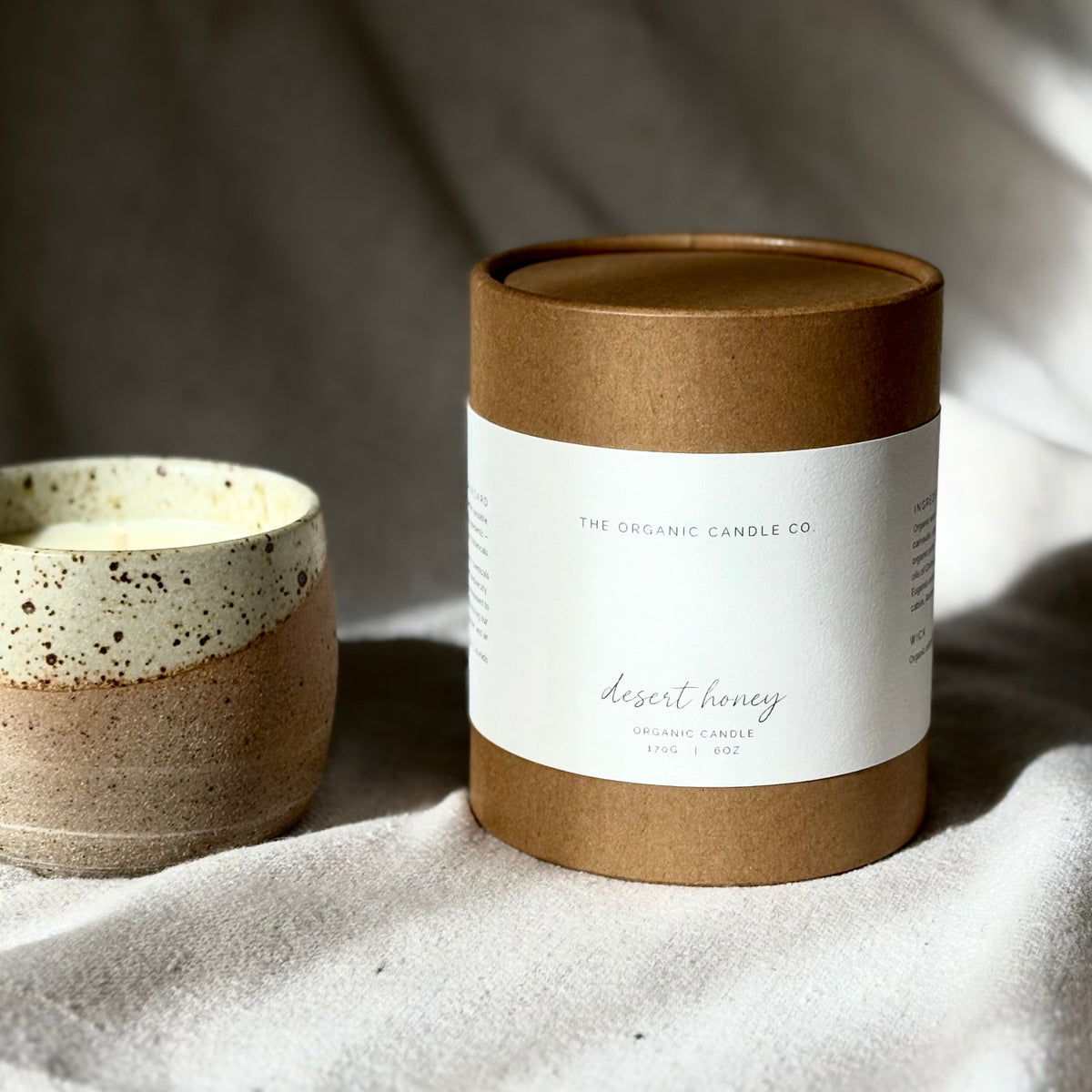 A cylindrical kraft paper candle tin with a white label reading 'THE ORGANIC CANDLE CO. desert honey' sits on draped fabric beside a speckled ceramic candle jar.