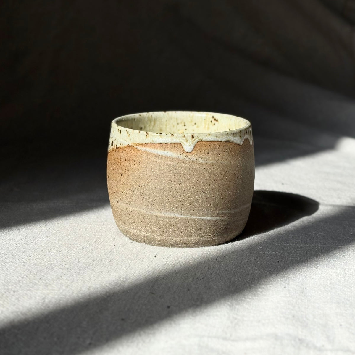 An organic candle in a handmade ceramic cup with a speckled beige and brown glaze and a contrasting cream-colored dripping rim, set on a textured light gray surface under dramatic natural sunlight and shadows.