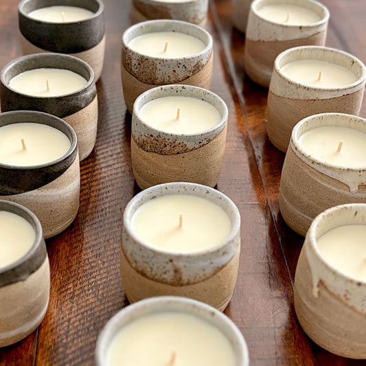 Multiple organic candles are arranged on a dark wooden surface. The candles are housed in rustic, speckled stoneware cups with a glossy dark rim.