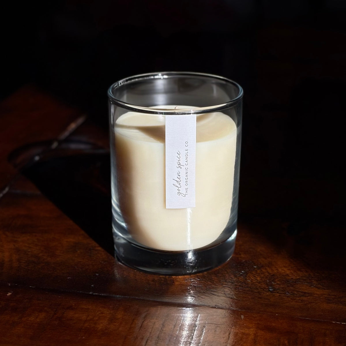 A clear glass organic candle filled with sits on a dark wooden table against a dark background. A small white label on the front reads 'golden spice' and 'The Organic Candle Co.