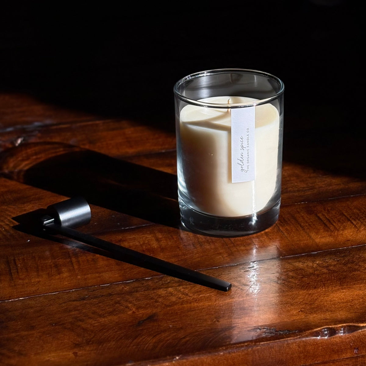 A clear glass organic candle fsits on a dark wooden table against a dark background. A small white label on the front reads 'golden spice' and 'The Organic Candle Co. A black metal wick snuffer is placed beside it in dramatic side lighting.