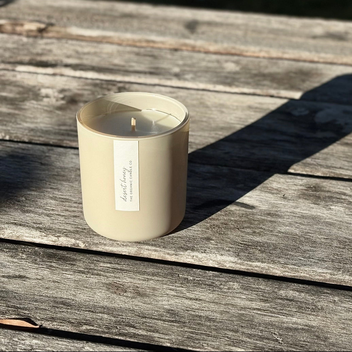 An organic candle in a beige-colored matte glass jar sits on a weathered wooden surface outdoors.