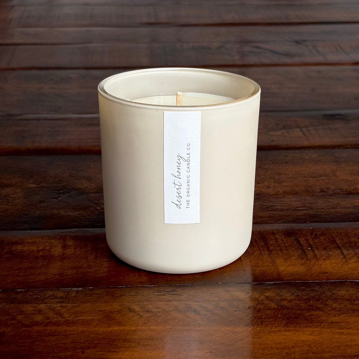An organic candle in a matte cream-colored jar sits on a dark wooden table, with a white label reading 'THE ORGANIC CANDLE CO.'