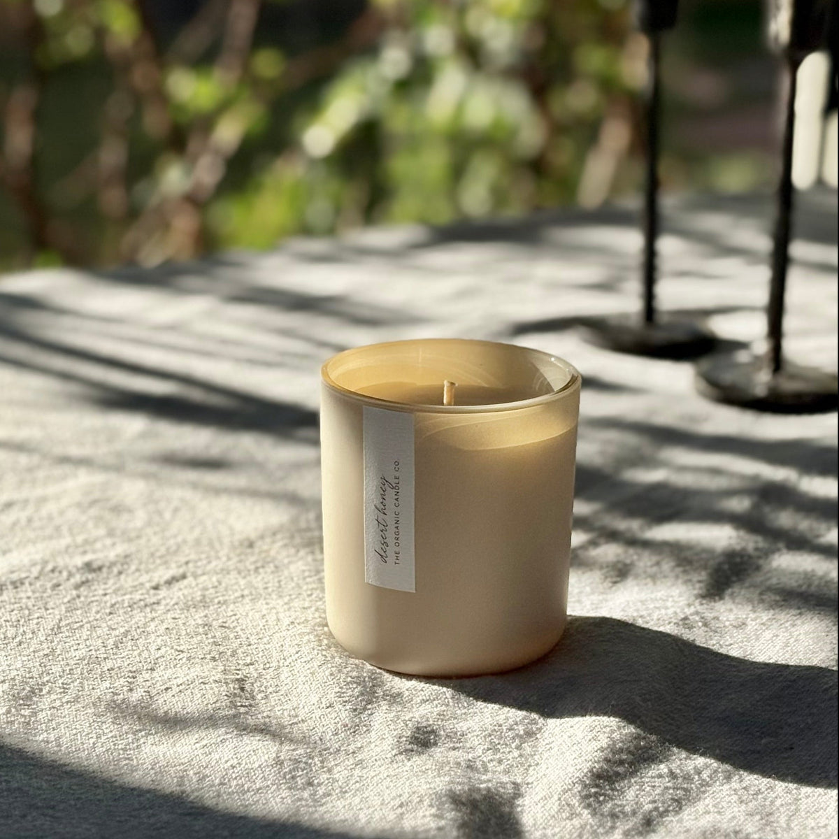 An organic candle in a beige-colored matte glass jar sits on a textured gray linen cloth outdoors, with dappled sunlight and soft green foliage blurred in the background.