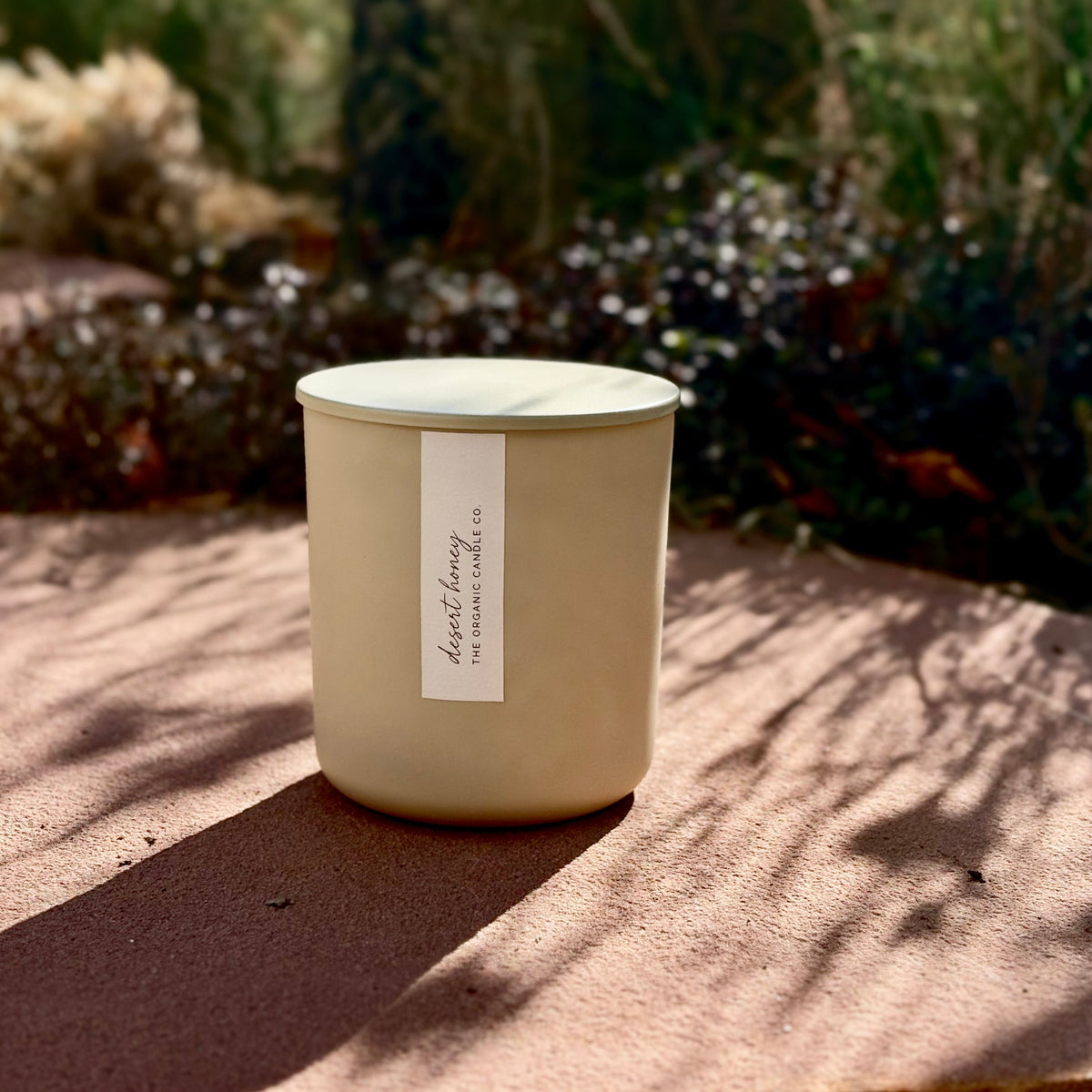 An organic candle in beige glass candle jar with a matching lid sits on a textured stone surface outdoors, with dappled sunlight and a blurred garden background. The front of the jar has a white label reading 'Desert Honey' and 'The Organic Candle Co.