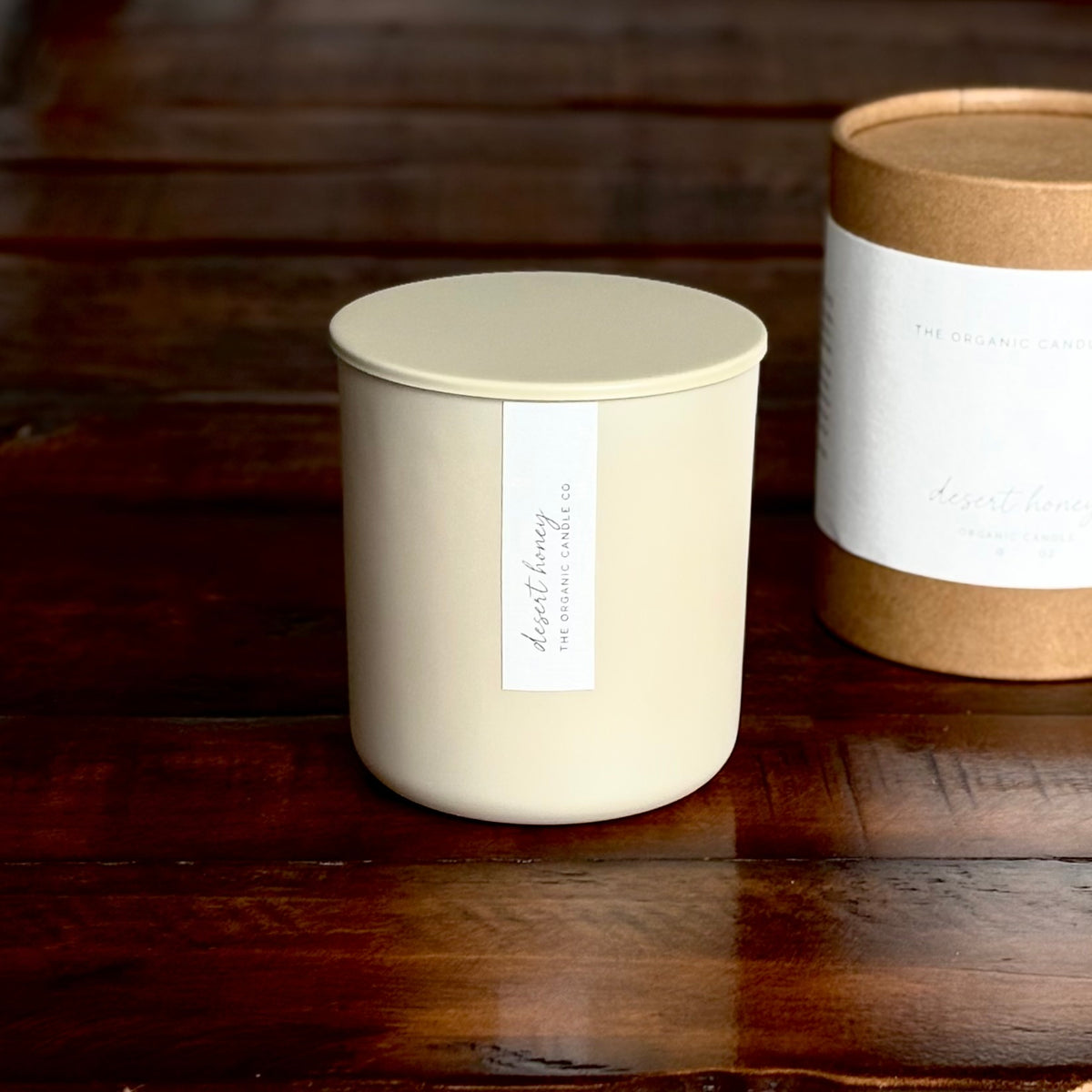 An organic candle in a cream-colored glass jar candle with a matching lid sits on a dark wooden surface, with its brown cardboard packaging cylinder partially visible in the background.